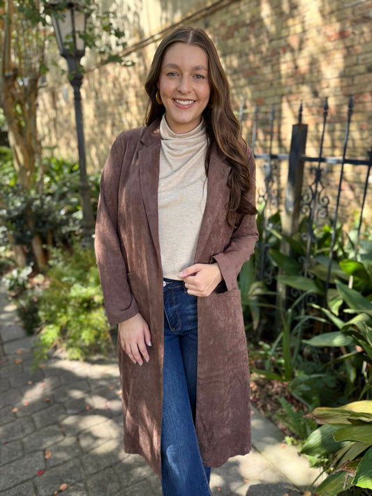 Woman wearing a long brown coat in an outdoor setting with plants and a brick wall.