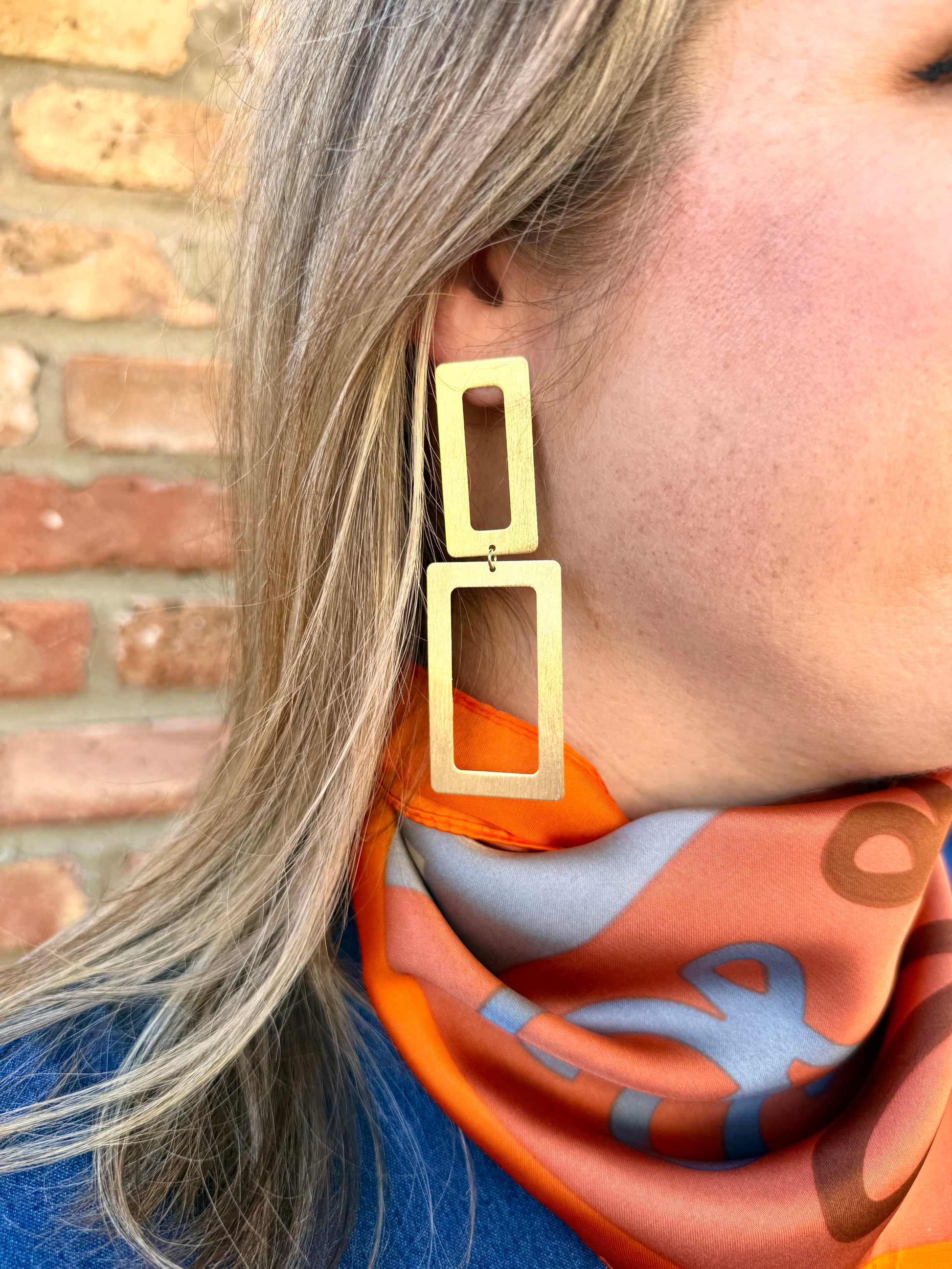 Gold hoop earrings worn by a person with a colorful scarf against a brick wall.
