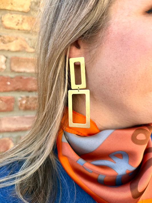 Gold hoop earrings worn by a person with a colorful scarf against a brick wall.