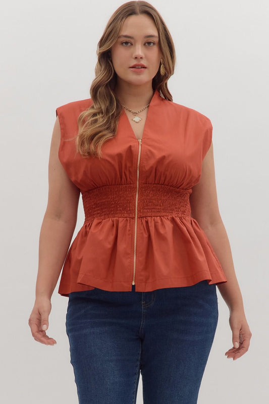 Woman wearing a rust-colored sleeveless top with a peplum waist against a plain background