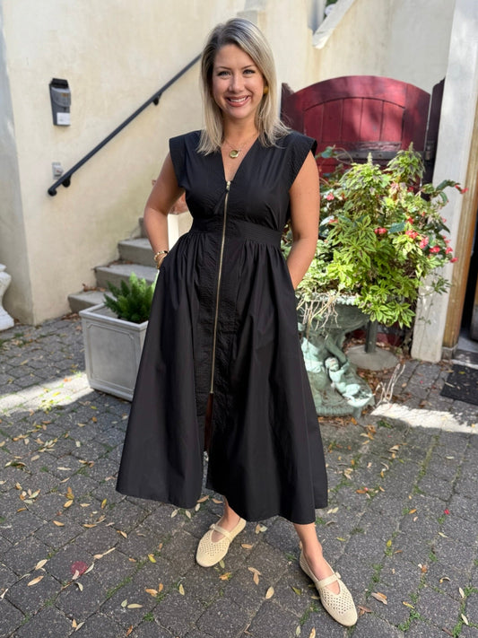 Woman in a black dress standing outdoors on a paved area with plants and steps in the background.